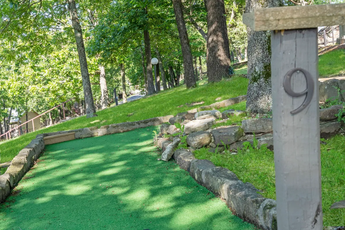 A vibrant outdoor mini-golf putting green nestled among trees, with a stone border and a numbered post marking the hole. The scene conveys a playful, family-friendly environment perfect for friendly competition and bonding.