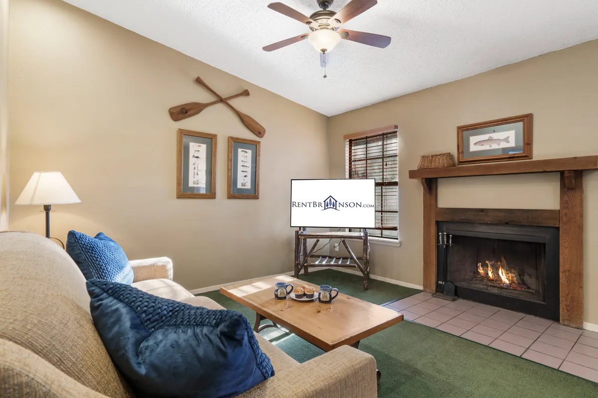 The image showcases a warm, inviting living room inside the condo. A plush sofa faces a crackling fireplace, with a coffee table set for games or snacks. Soft lighting and tasteful wall decor create a cozy, home-like ambiance, while a television sits ready for movie nights. Large windows hint at tranquil wooded views outside, enhancing the sense of peaceful retreat.