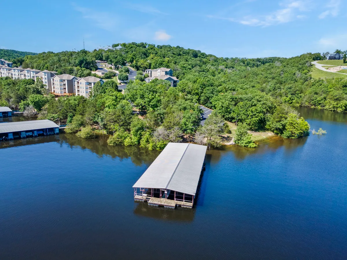 This image captures Table Rock Lake with a boathouse prominently featured, surrounded by lush greenery and apartments perched on a nearby hill. The tranquil water reflects the peaceful surroundings, and the dock offers easy access for boating and fishing.