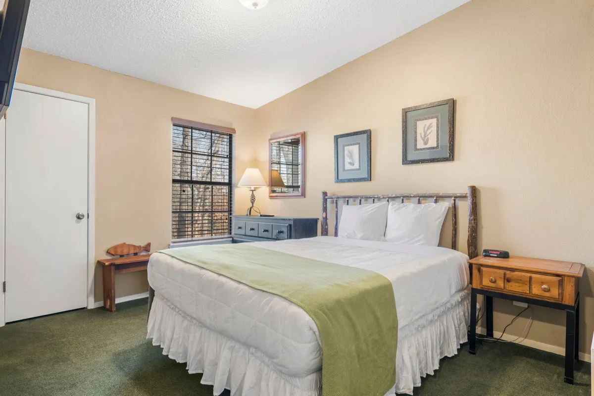 A tranquil bedroom featuring a queen bed dressed in white linens and a green throw blanket, flanked by wooden nightstands and soft lamps. Framed artwork above the bed and ample natural light from a nearby window create a serene, restful setting.