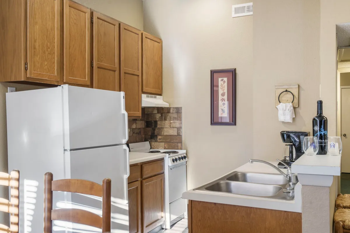 Warm kitchen scene with a white refrigerator, wooden cabinets, a stainless steel sink, and various kitchen appliances. The space is designed for cooking and social gathering, evoking the comfort of home-cooked meals.