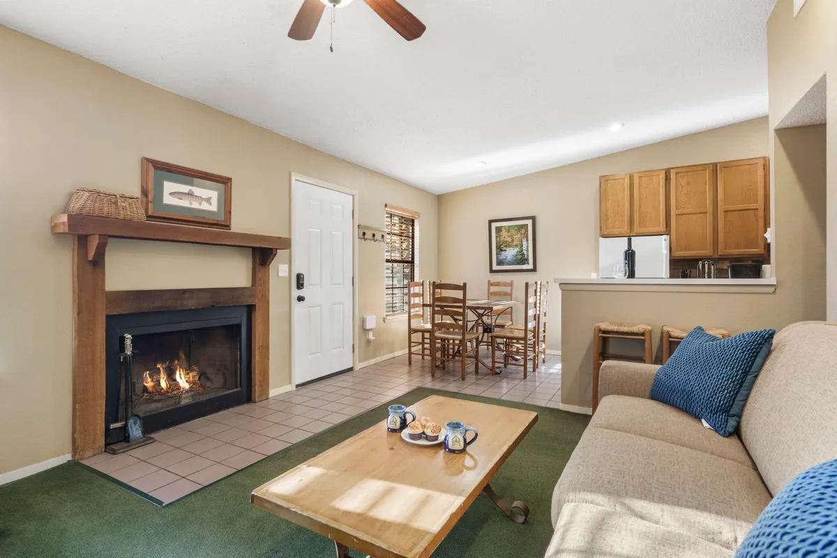 A wide shot of the open-concept living area featuring a plush sofa, a stone-clad fireplace, a wooden dining table set for meals, and a glimpse of the kitchen beyond. Warm natural light streams in through the windows, highlighting the cozy textures and inviting atmosphere.