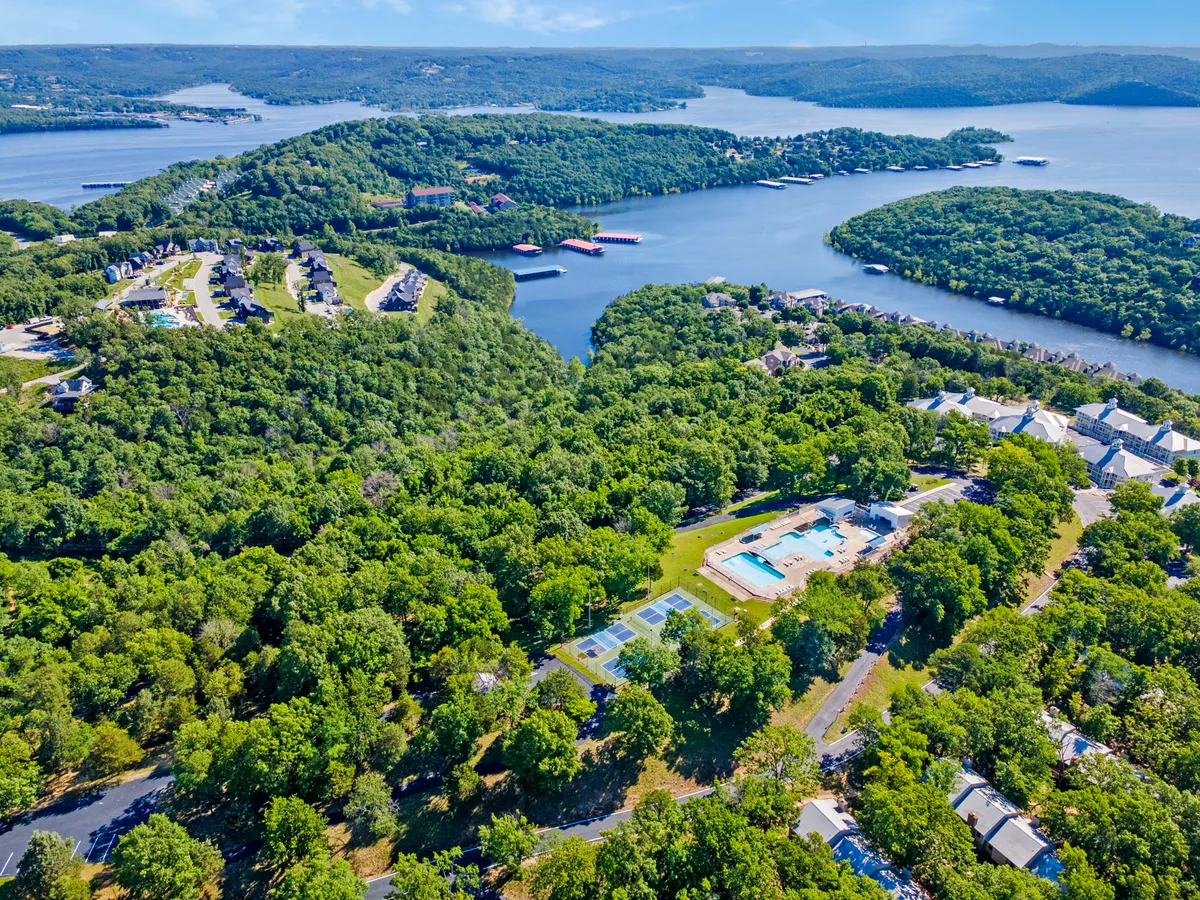 An exterior shot featuring inviting swimming pools, tennis courts, and lush grounds with a view of the lake and residential buildings. The scene highlights multiple resort amenities and the relaxing, recreational environment available to guests.
