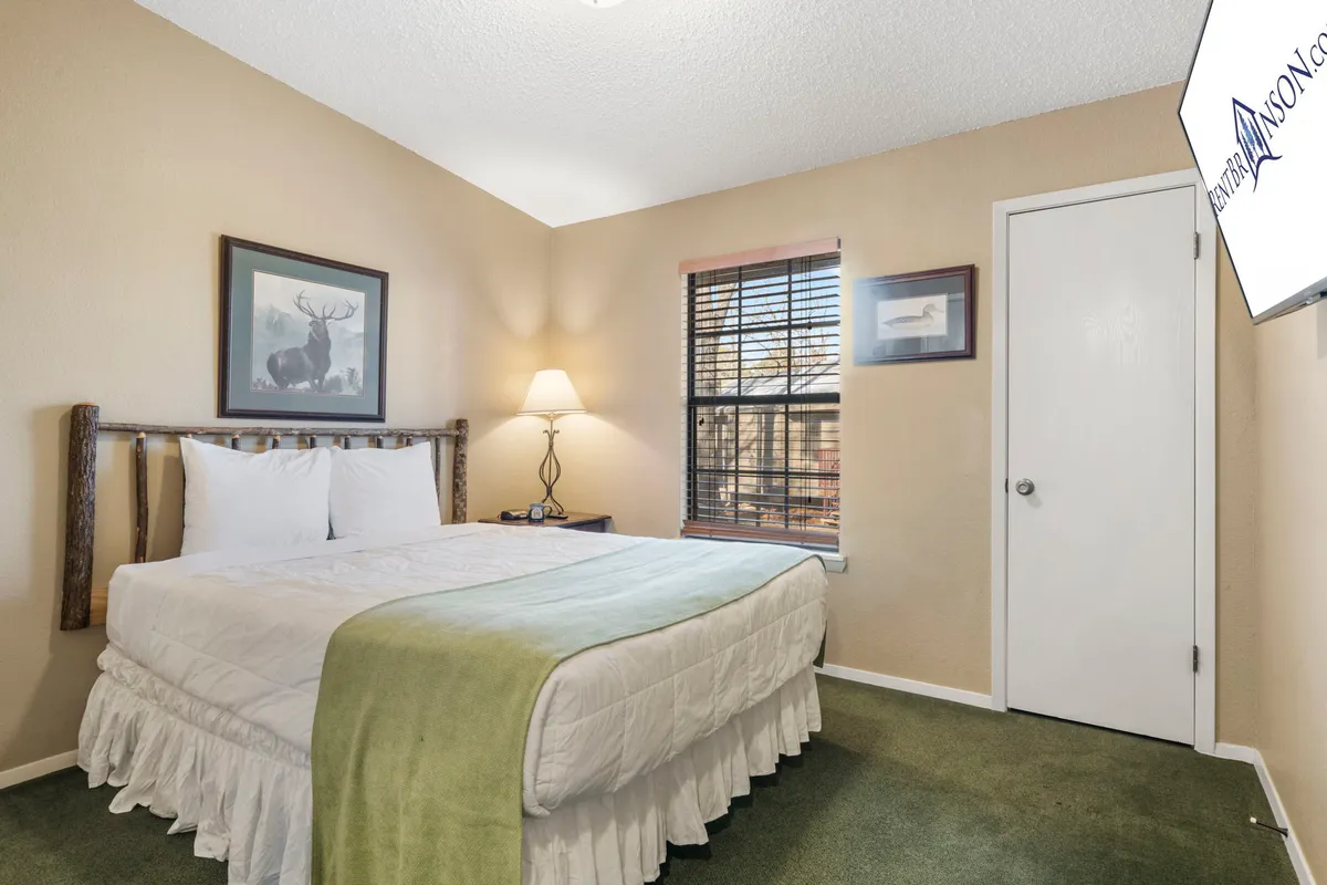 A peaceful bedroom with a plush queen bed adorned with white linens and a green throw blanket. The room includes a table lamp, framed artwork, and a window with blinds allowing soft natural light to filter in, highlighting the relaxing mood.