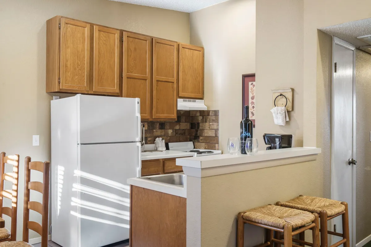 A cozy, functional kitchen space featuring kitchen cabinets, a white refrigerator, bar stools at a breakfast counter, and visible kitchen appliances. The layout highlights ample counter space and seating for casual meals or gatherings.